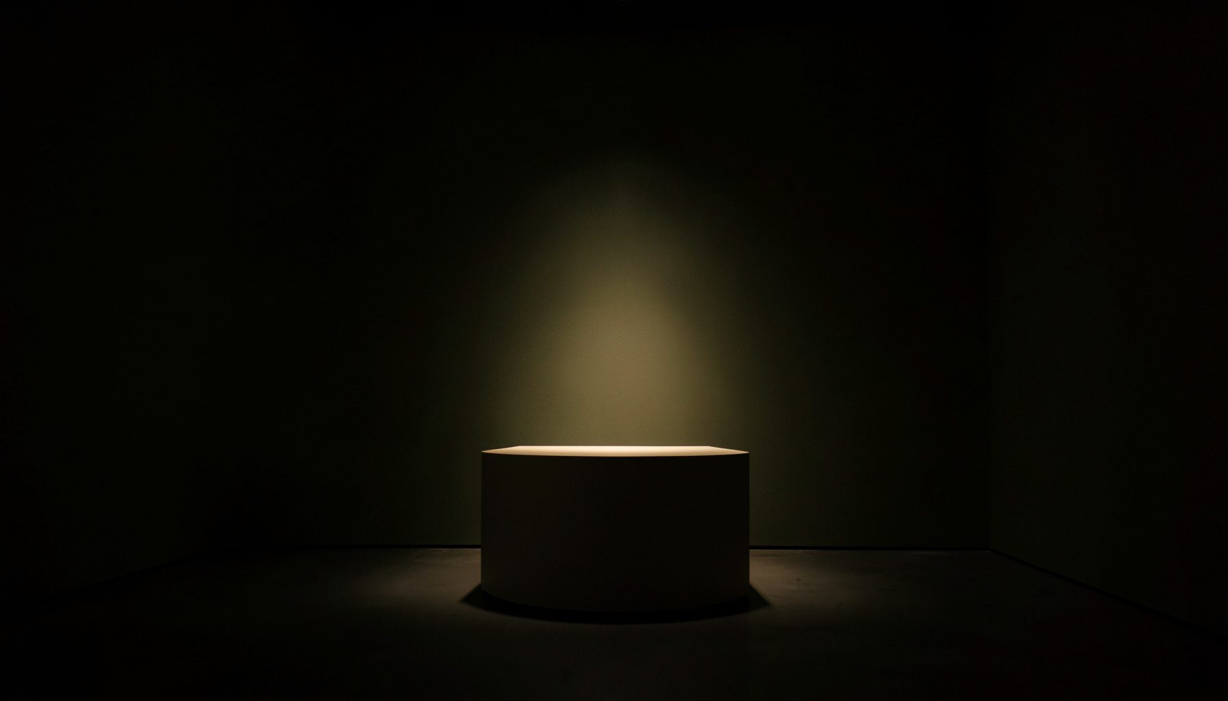 A dark, low-key photograph of a quiet gallery interior with deep shadow, muted olive walls, and a single warm spotlight